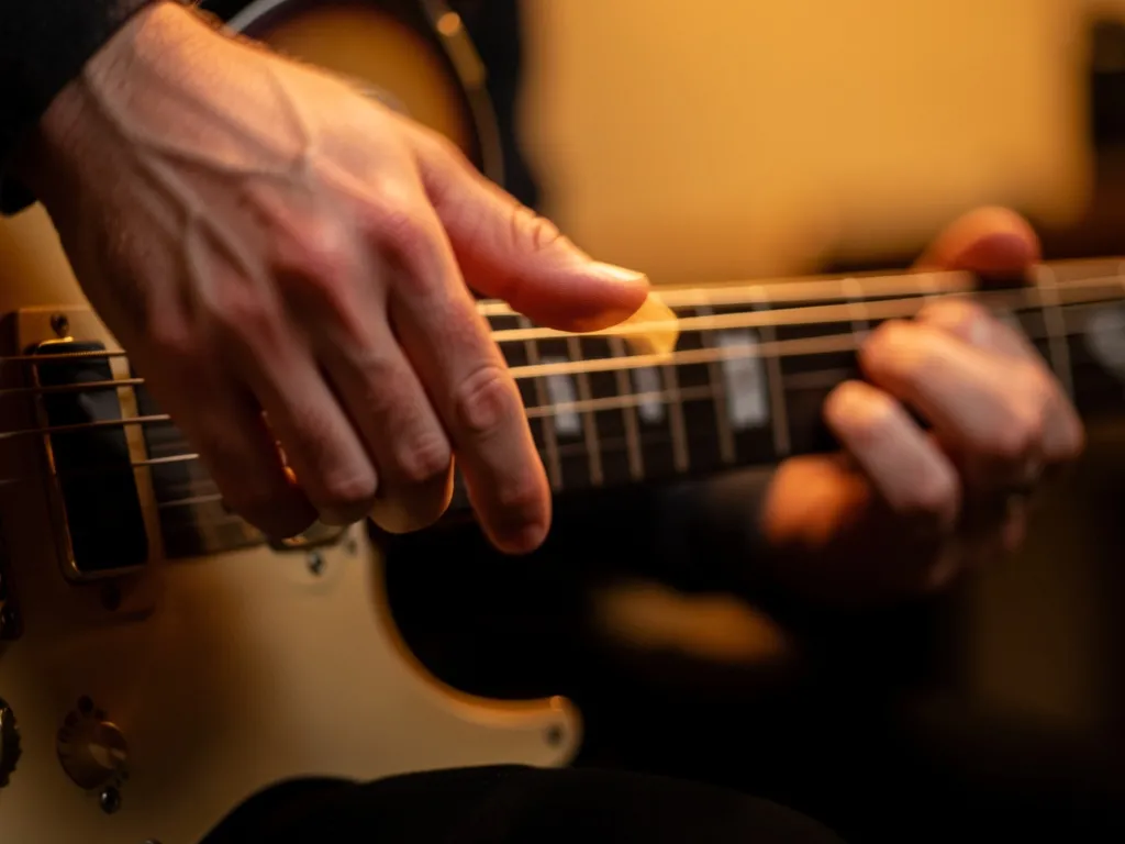 Close-up da mão esquerda de um guitarrista executando um hammer-on no braço da guitarra com iluminação dramática e desfoque de movimento